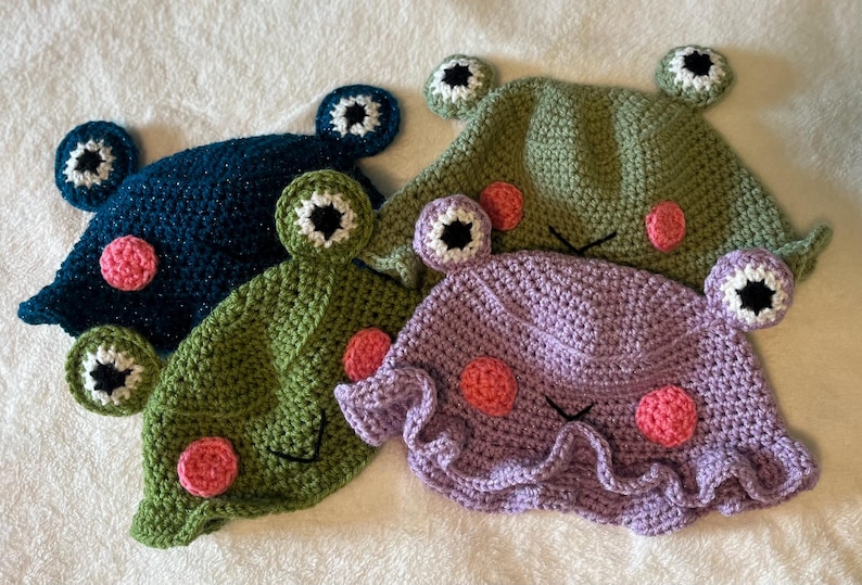 May include: Four handmade crochet frog hats in shades of blue, green, and purple. Each hat features large, round eyes with white and black details, pink cheek circles, and a stitched mouth. The hats are designed to be worn.