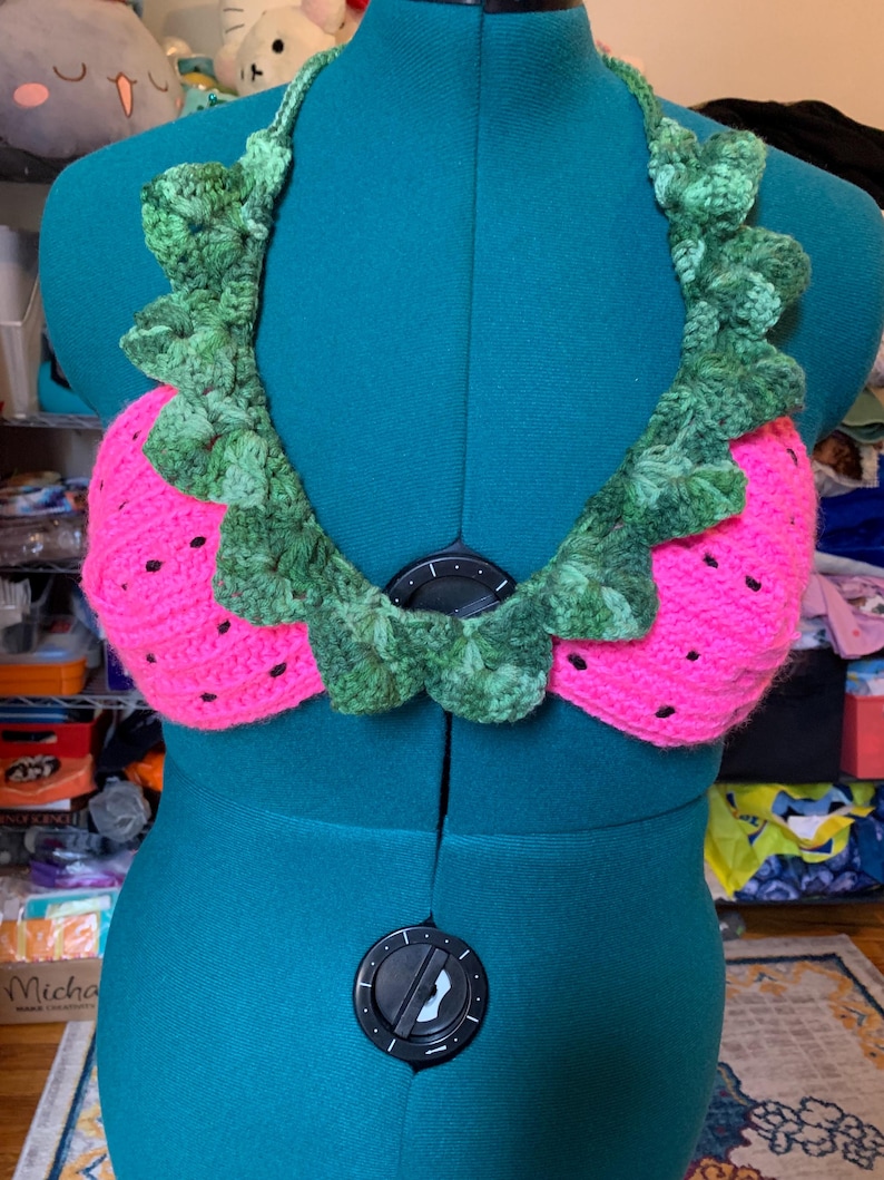 May include: A handmade crochet top in the shape of a watermelon. The top features a bright pink base with black seed details and a green leafy trim around the neckline. The halter top is displayed on a teal mannequin.