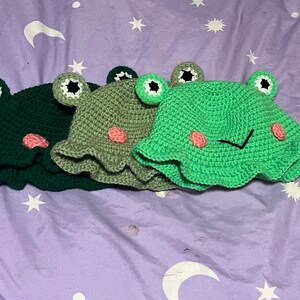 May include: Three handmade crochet frog hats in shades of green. Each hat features large, round eyes with black pupils and pink cheeks. The hats have a ruffled brim and are designed to resemble a frog's face. The hats are displayed on a purple surface.