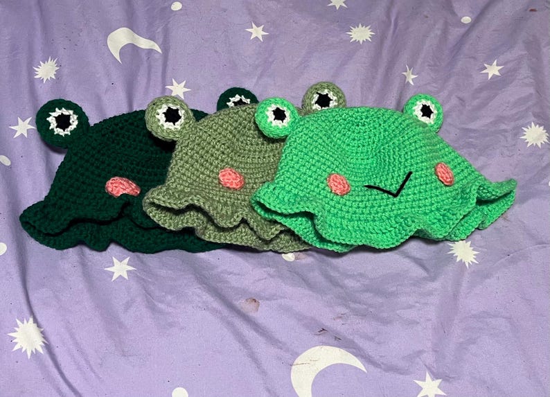 May include: Three handmade crochet frog hats in shades of green. Each hat features large, round eyes with black pupils and pink cheeks. The hats have a ruffled brim and are designed to resemble a frog's face. The hats are displayed on a purple surface.