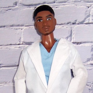 May include: A doll wearing a white lab coat over a light blue scrub top. The doll has short black hair and brown eyes.