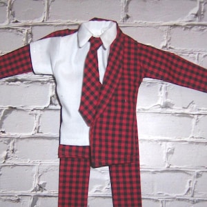 May include: A doll-sized outfit featuring a white collared shirt, a red and black plaid blazer, a matching tie, and plaid pants. The miniature suit is displayed against a white brick wall.