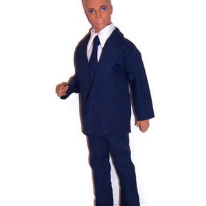 May include: A doll wearing a dark blue suit with a white shirt and a dark blue tie. The doll has short blond hair and is wearing black shoes.