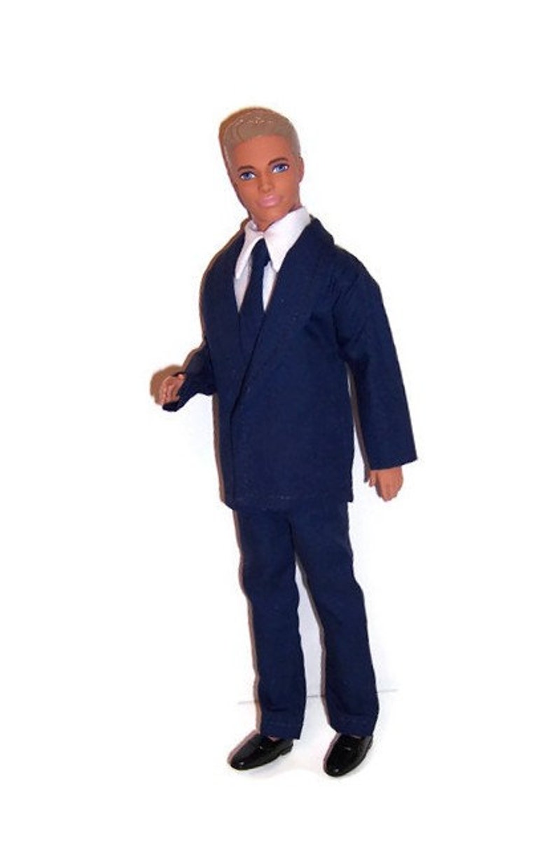 May include: A doll wearing a dark blue suit with a white shirt and a dark blue tie. The doll has short blond hair and is wearing black shoes.