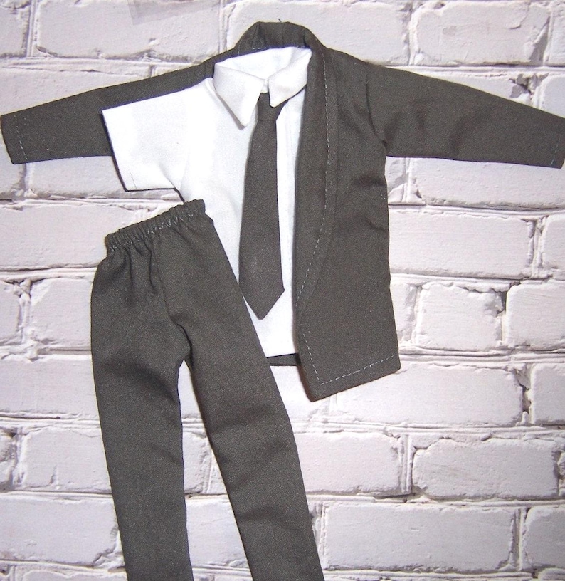 K&ouml;nnte beinhalten: Ein dunkelgrauer Anzug mit lang&auml;rmeliger Jacke, wei&szlig;em Hemd mit Kragen, dunkelgrauer Krawatte und passender Hose mit elastischem Bund. Das Set ist als Miniatur-Outfit f&uuml;r eine Puppe oder eine kleine Figur konzipiert.