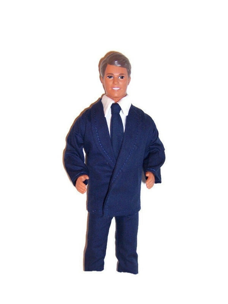 May include: A doll wearing a dark blue suit with a white shirt and a dark blue tie.