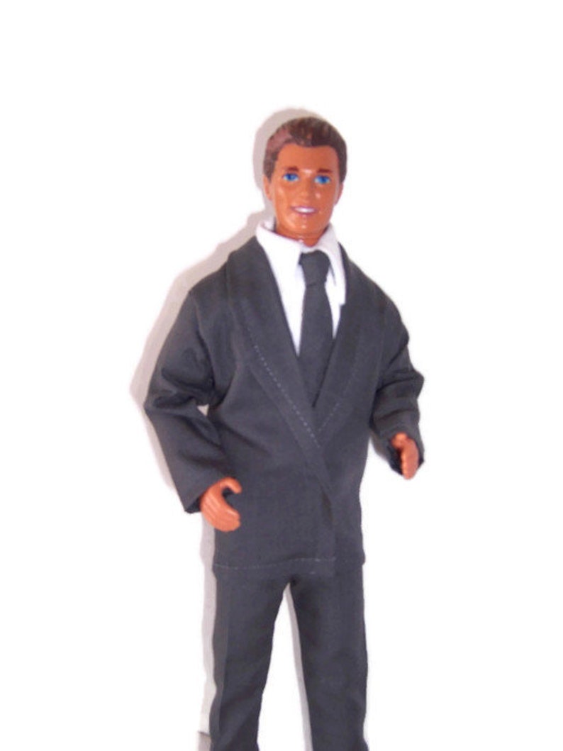 K&ouml;nnte beinhalten: Eine Kunststoffpuppe mit braunen Haaren und blauen Augen, bekleidet mit einem dunkelgrauen Sakko, einer passenden Hose, einem wei&szlig;en Hemd mit Kragen und einer dunkelgrauen Krawatte. Die Puppe hat einen neutralen Gesichtsausdruck und steht aufrecht.