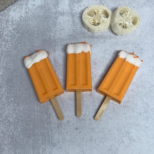 May include: Three orange popsicle-shaped soaps with white tops and wooden sticks. The soaps are arranged in a triangular pattern on a gray background.