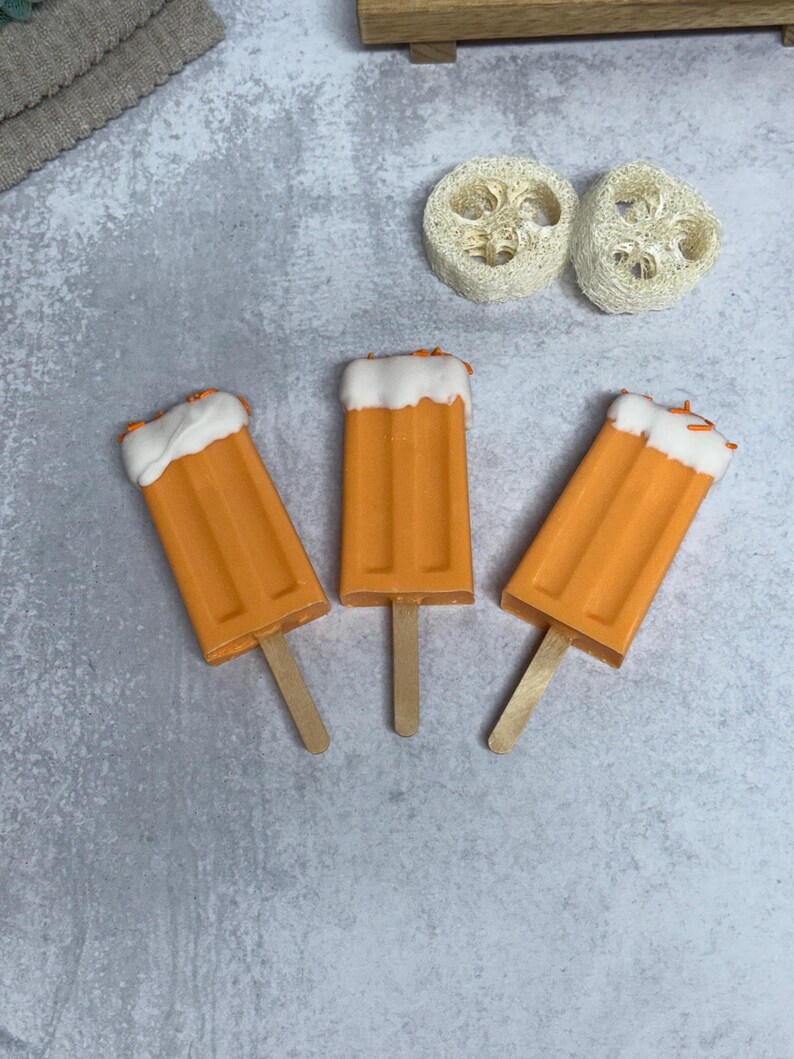 May include: Three orange popsicle-shaped soaps with white tops and wooden sticks. The soaps are arranged in a triangular pattern on a gray background.