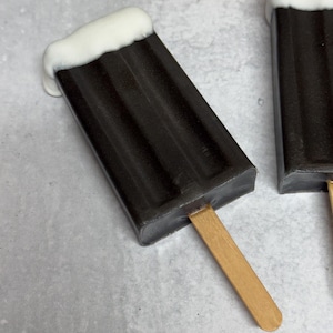 May include: Two chocolate popsicles with white chocolate topping on a stick. The popsicles are on a gray surface.