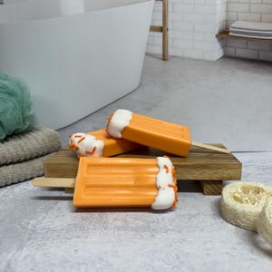 May include: Three orange and white popsicle shaped soaps with orange sprinkles on a wooden tray. The soaps are in a bathroom setting.