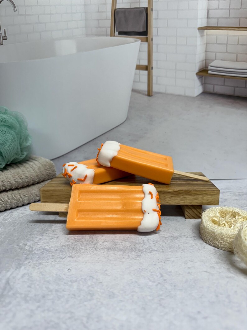 May include: Three orange and white popsicle shaped soaps with orange sprinkles on a wooden tray. The soaps are in a bathroom setting.