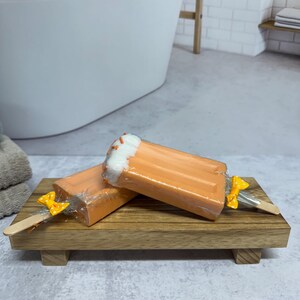 May include: Two orange and white popsicle shaped soaps on a wooden tray. The soaps have a yellow bow on a wooden stick.