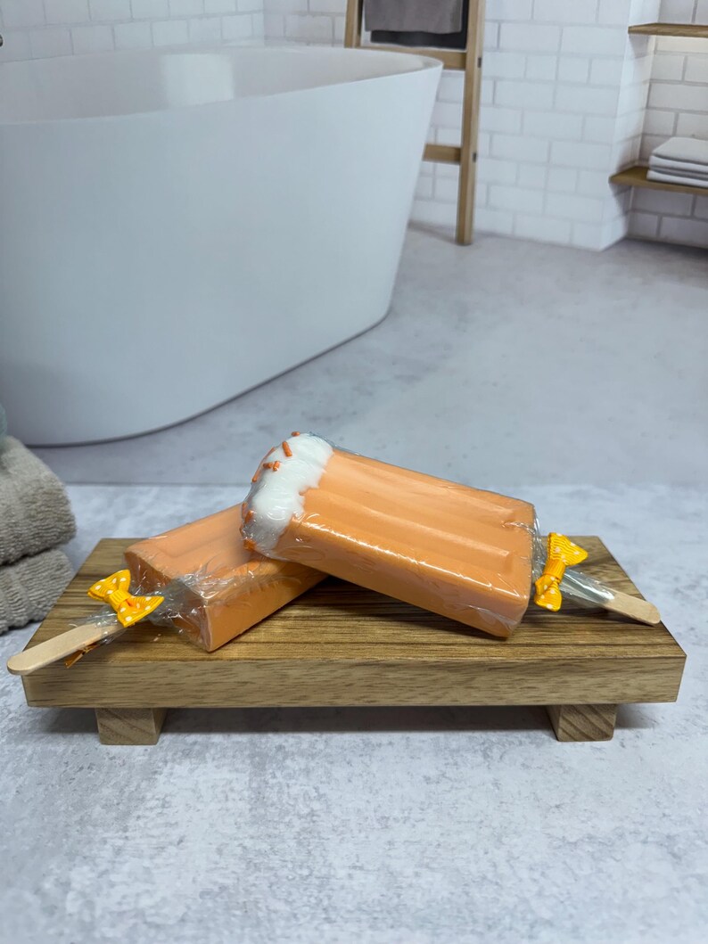 May include: Two orange and white popsicle shaped soaps on a wooden tray. The soaps have a yellow bow on a wooden stick.