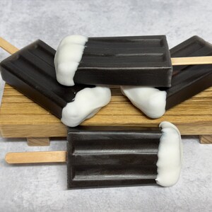 May include: Four chocolate and white popsicle soaps stacked on a wooden tray. The soaps are shaped like popsicles with a white top and a dark chocolate bottom.