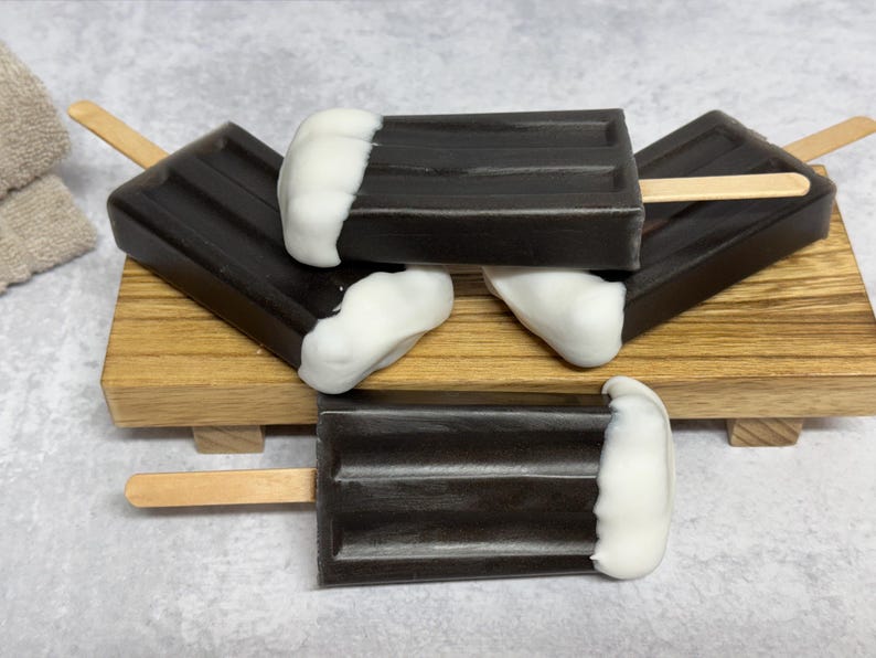 May include: Four chocolate and white popsicle soaps stacked on a wooden tray. The soaps are shaped like popsicles with a white top and a dark chocolate bottom.