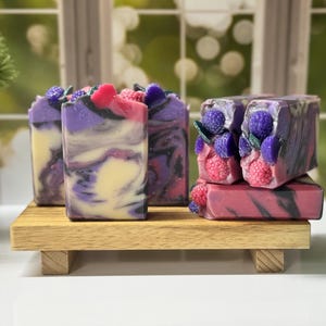 May include: Artisan soaps with a berry theme. The soaps are swirled with purple, pink, and cream colors. Some soaps are topped with decorative purple berries and pink raspberries. The soaps are displayed on a small wooden tray.