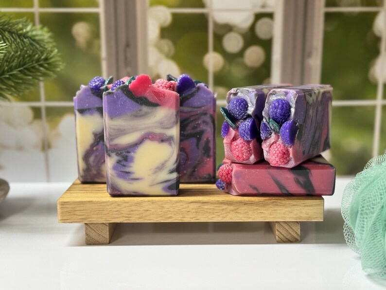 May include: Artisan soaps with a berry theme. The soaps are swirled with purple, pink, and cream colors. Some soaps are topped with decorative purple berries and pink raspberries. The soaps are displayed on a small wooden tray.