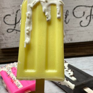 May include: A yellow popsicle with white chocolate and sprinkles on top. The popsicle is on a wooden stick and is being held by a hand.