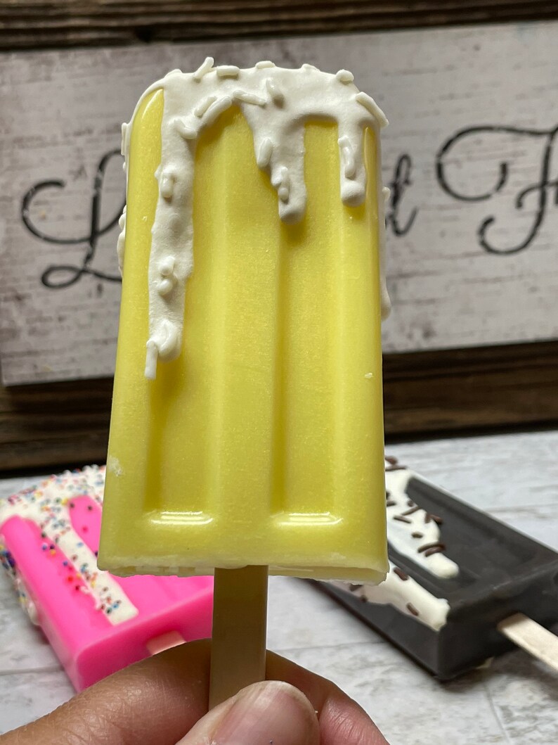 May include: A yellow popsicle with white chocolate and sprinkles on top. The popsicle is on a wooden stick and is being held by a hand.