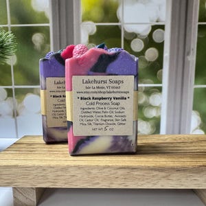 May include: Two bars of handmade soap with a layered design of purple, pink, and white, topped with decorative elements. Each bar has a label with the text "Black Raspberry Vanilla" and "Cold Process Soap". The soaps are displayed on a wooden tray.