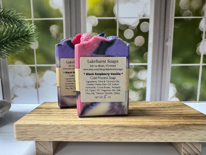 May include: Two bars of handmade soap with a layered design of purple, pink, and white, topped with decorative elements. Each bar has a label with the text "Black Raspberry Vanilla" and "Cold Process Soap". The soaps are displayed on a wooden tray.