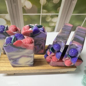 May include: Artisan soaps with a berry theme. The soaps are swirled with purple, pink, and white colors, and topped with decorative berries. The soaps are displayed on a small wooden tray. The background is a window.