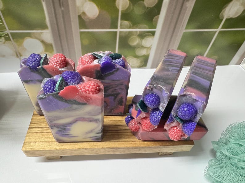 May include: Artisan soaps with a berry theme. The soaps are swirled with purple, pink, and white colors, and topped with decorative berries. The soaps are displayed on a small wooden tray. The background is a window.