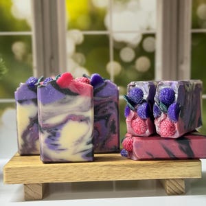 May include: Artisan soaps with a swirl design in shades of purple, black, and cream, topped with decorative berries. The soaps are displayed on a small wooden shelf, creating a natural and elegant presentation.