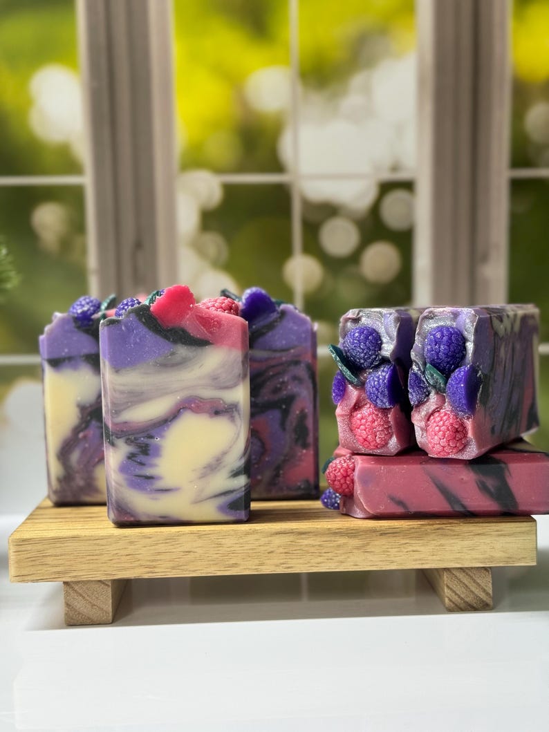May include: Artisan soaps with a swirl design in shades of purple, black, and cream, topped with decorative berries. The soaps are displayed on a small wooden shelf, creating a natural and elegant presentation.