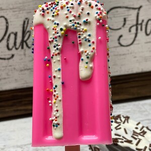 May include: A pink popsicle with white chocolate and colorful sprinkles. The popsicle is on a wooden stick.