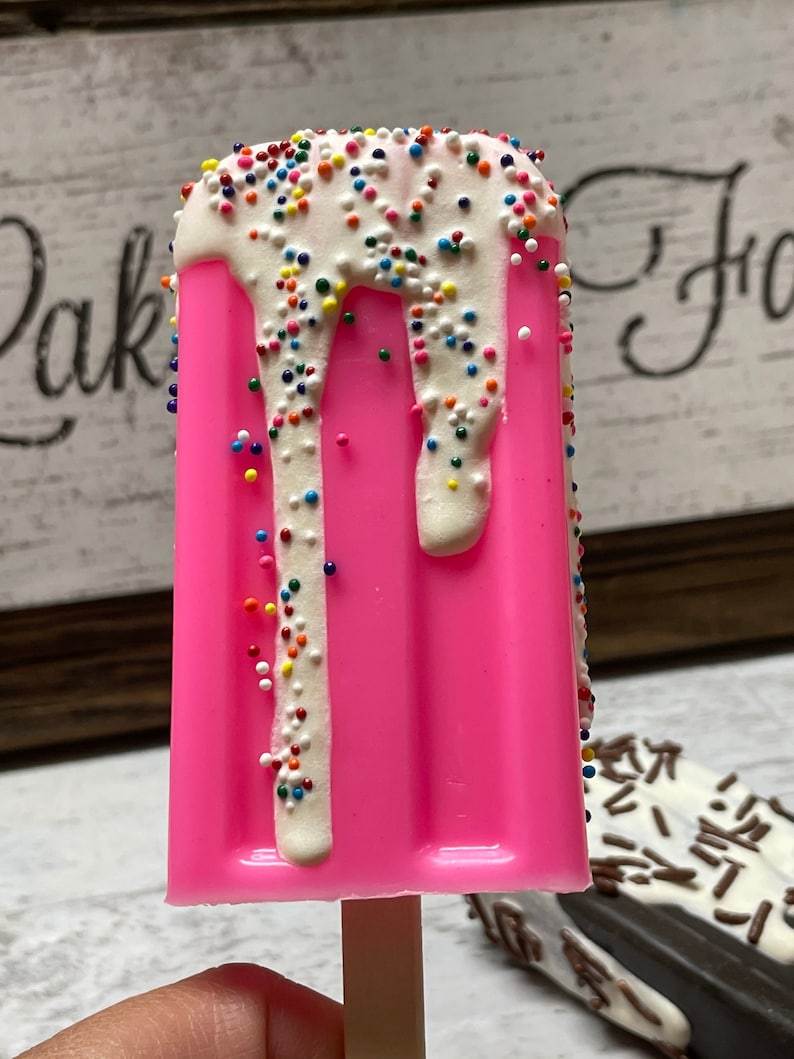 May include: A pink popsicle with white chocolate and colorful sprinkles. The popsicle is on a wooden stick.