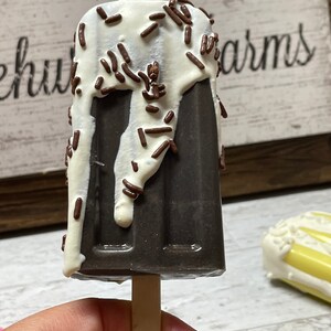May include: A chocolate popsicle with white chocolate drizzle and chocolate sprinkles on a wooden stick. The popsicle is held in a hand.