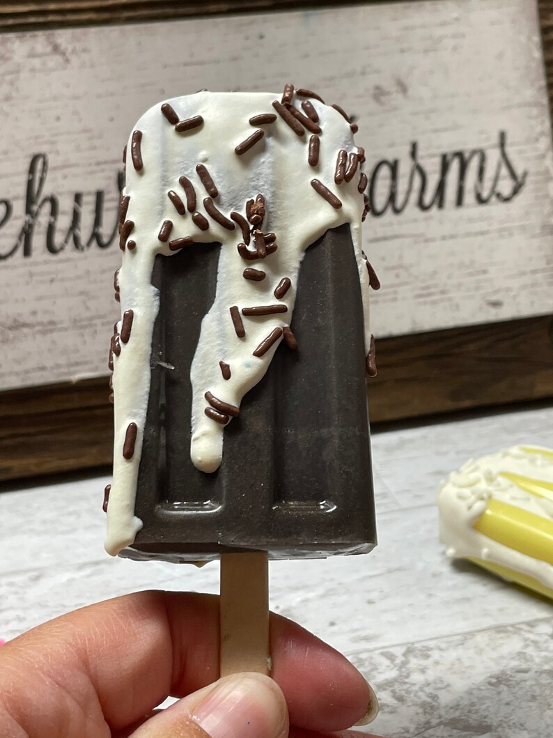 May include: A chocolate popsicle with white chocolate drizzle and chocolate sprinkles on a wooden stick. The popsicle is held in a hand.