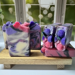May include: Artisan soaps with a berry theme. The soaps are rectangular, with a marbled appearance in shades of purple, white, and pink. Some soaps are topped with decorative berries and leaves, and are displayed on a small wooden tray.