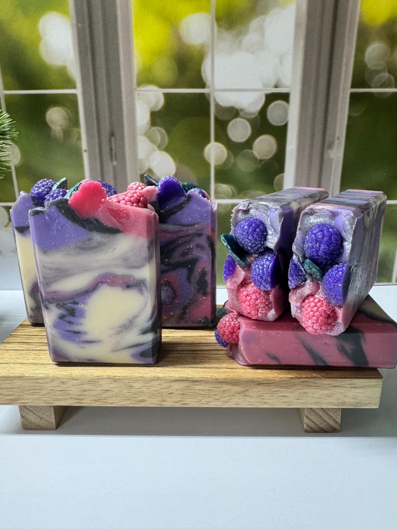 May include: Artisan soaps with a berry theme. The soaps are rectangular, with a marbled appearance in shades of purple, white, and pink. Some soaps are topped with decorative berries and leaves, and are displayed on a small wooden tray.