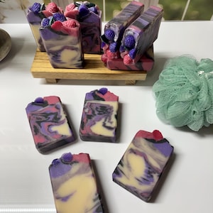 May include: Artisan soaps with a swirled design in shades of purple, pink, and cream. Some soaps are topped with decorative berry-shaped embellishments. The soaps are arranged on a wooden tray and a white surface.