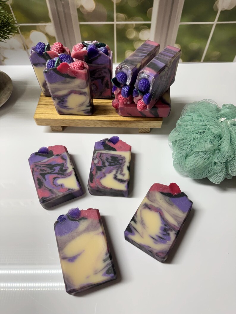 May include: Artisan soaps with a swirled design in shades of purple, pink, and cream. Some soaps are topped with decorative berry-shaped embellishments. The soaps are arranged on a wooden tray and a white surface.