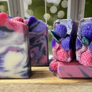 May include: Artisan soap bars in shades of purple, pink, and white, with decorative raspberry and blueberry accents. The soaps have a marbled appearance and are arranged on a wooden surface. The soaps are handmade and have a natural aesthetic.
