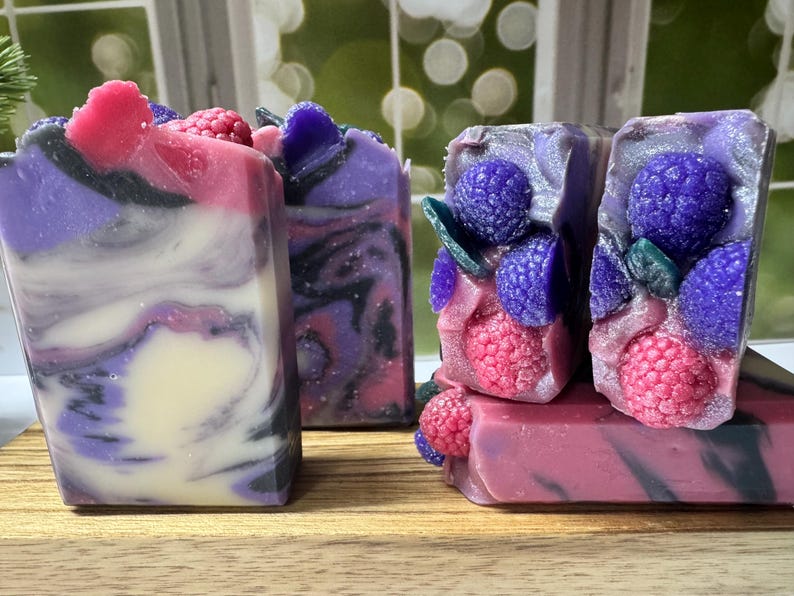 May include: Artisan soap bars in shades of purple, pink, and white, with decorative raspberry and blueberry accents. The soaps have a marbled appearance and are arranged on a wooden surface. The soaps are handmade and have a natural aesthetic.