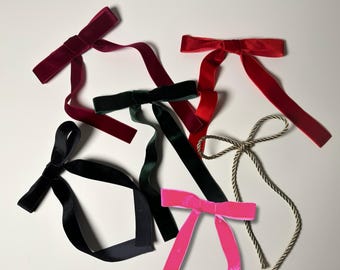 May include: Six velvet ribbon bows in various colors, including red, black, green, and pink. The bows are tied with long tails and are arranged on a white surface.