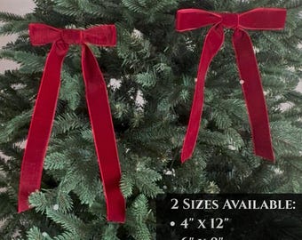 May include: Two red velvet bows with long tails are displayed on a green Christmas tree. The bows are a deep red color and have a soft, velvety texture. The image includes text that says "2 SIZES AVAILABLE: 4" x 12" and 6" x 8".