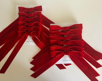 May include: Two sets of red velvet bows with long, flowing tails. Each set has five bows attached to a white card. The text "KIND+BRIGHT" is printed on the cards. The bows are a deep, rich red color.