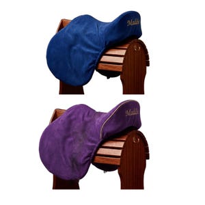 Op de afbeelding: Twee zadelhoezen, één in donkerblauw en één in paars, elk met de naam Maddie geborduurd in gouddraad aan de zijkant. Deze zachte stoffen hoezen zijn ontworpen om over een zadel te passen en worden getoond op houten zadelsteunen.