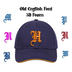 Op de afbeelding: Een marineblauwe baseballpet met een oranje Old English letter "H" geborduurd op de voorkant. De pet heeft een gele rand en de tekst "Old English Font 3D Foam" erboven. Extra Old English letters in blauw, roze en rood omringen de pet.