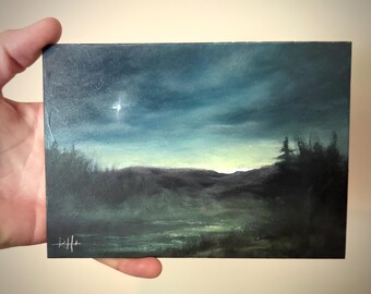 May include: An original landscape painting depicting a night scene with a bright star in the sky. The artwork features dark blues and greens, with a hint of yellow at the horizon. The painting is held by a hand.