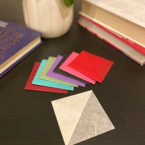 May include: A set of eight colorful paper bookmarks, folded into triangles, with a white and gray paper bookmark in the foreground.