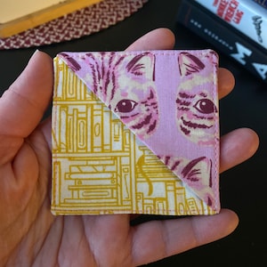May include: A pink and yellow fabric bookmark with a cat face on one side and a pattern of books on the other side.