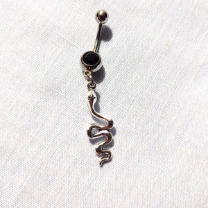 May include: A silver belly button ring with a black gem and a dangling snake charm. The curved barbell has a small silver ball on top. The snake charm is detailed and coiled.