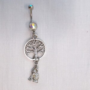 May include: Silver-toned belly button ring with a curved barbell. The top and center feature iridescent gems. A circular tree of life charm and a howling wolf pendant dangle below, creating a nature-inspired design.